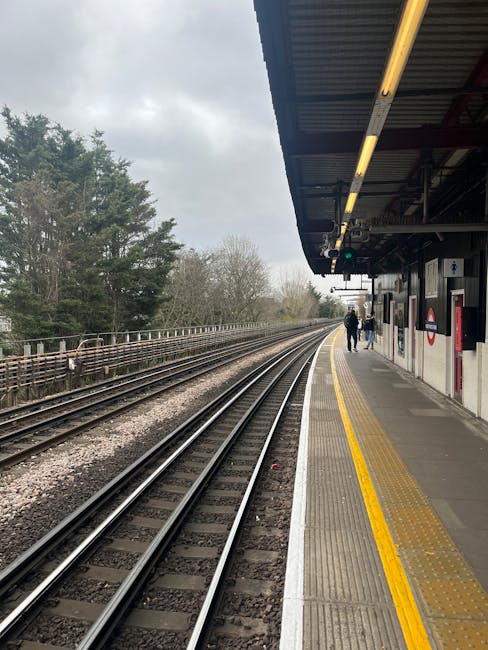 A view of Colliers Wood Tube Station platform with train tracks running parallel to the platform edge, which has a textured yellow safety strip along its length. The platform is covered by a dark meta