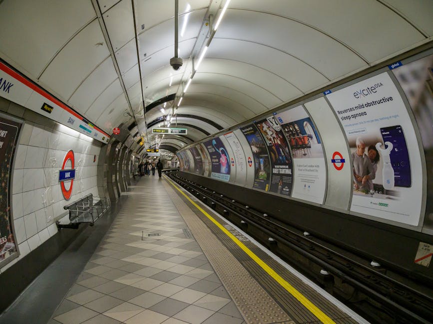 A view of Colliers Wood Tube Station platform with train tracks running parallel to the platform edge, which has a textured yellow safety strip along its length. The platform is covered by a dark meta